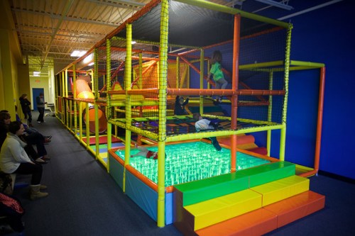 A colorful indoor play structure with nets, slides, and climbing areas, filled with children playing and adults watching.