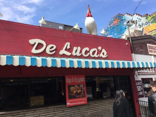 A red storefront with "De Luca's" sign, featuring a large chicken statue on the roof and people walking by.