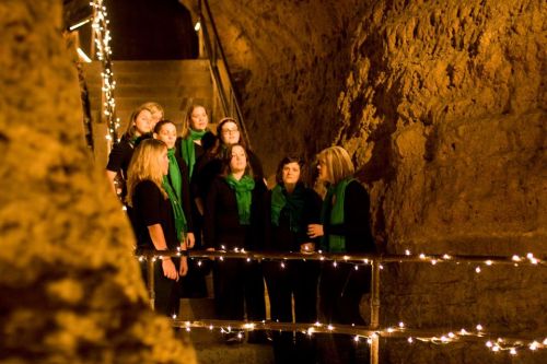 A choir of women in black outfits and green scarves sings on a staircase adorned with fairy lights in a cave-like setting.