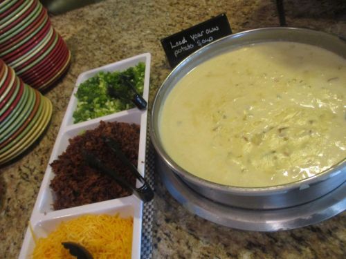 A bowl of creamy potato soup with toppings: chopped green onions, bacon bits, and shredded cheese on the side.