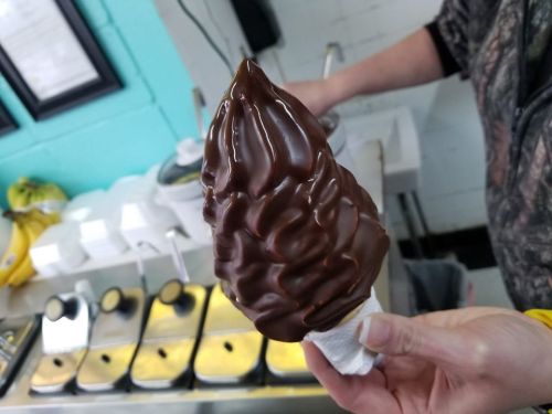 A hand holds a tall, swirled chocolate soft serve ice cream cone against a colorful background.