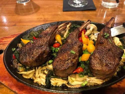 Sizzling platter of grilled lamb chops with colorful peppers and greens on a wooden table.