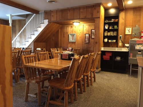Cozy wooden dining area with tables and chairs, featuring rustic decor and a staircase in the background.