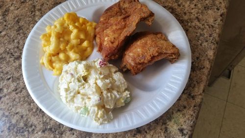 A paper plate with two pieces of fried chicken, macaroni and cheese, and a serving of potato salad.