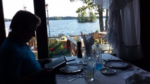 A person sits at a table by a lake, reading a menu, with a scenic view of water and trees in the background.
