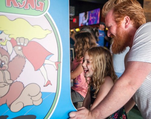 A man and a girl joyfully play an arcade game together, laughing in a lively gaming environment.