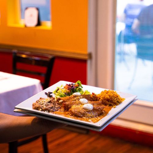 A person holds a plate with enchiladas, rice, beans, and a side salad, set against a colorful restaurant background.