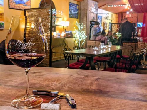 A glass of red wine on a wooden table, with a corkscrew nearby and a cozy restaurant ambiance in the background.