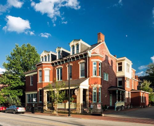 Historic red brick house with a porch, surrounded by trees, under a blue sky with scattered clouds.