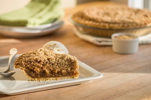 A slice of pie with a crumbly topping on a white plate, with a whole pie and ice cream in the background.