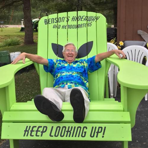 An elderly man in a colorful shirt sits joyfully in a large green chair with an alien design, smiling at the camera.
