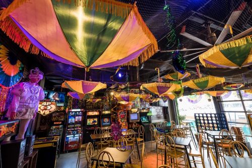Colorful umbrellas hang from the ceiling of a vibrant arcade, with tables and vintage arcade games visible below.