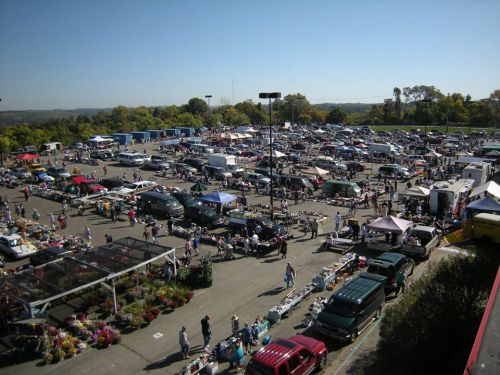 Aerial view of a bustling flea market with numerous vendors, cars, and shoppers under a clear blue sky.