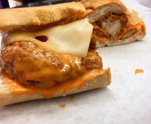 A close-up of a sandwich with crispy chicken, melted cheese, and spicy sauce on a toasted bun, resting on parchment paper.