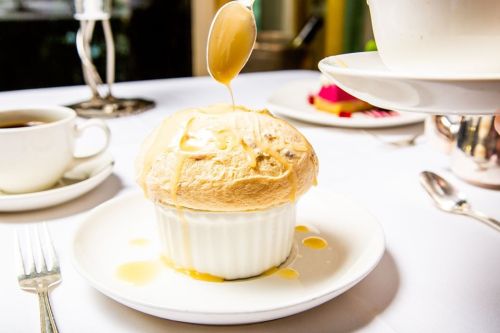 A fluffy dessert in a white ramekin, drizzled with sauce, sits on a plate beside a cup of coffee.