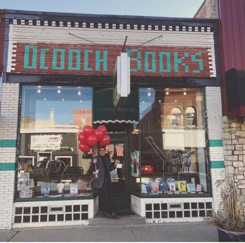 A person stands outside a bookstore holding red balloons, with books displayed in the window and a colorful storefront.