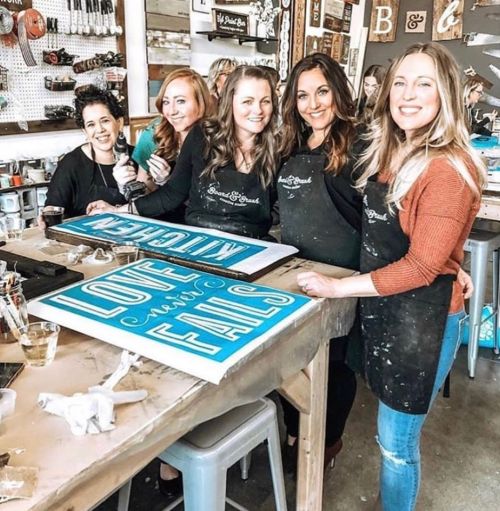 Five women in aprons smile while holding painted signs at a crafting workshop, surrounded by art supplies.