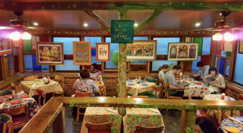 Cozy restaurant interior with colorful tablecloths, patrons dining, and artwork on the walls. A sign reads "Be Nice or Leave."