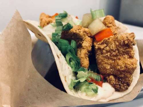 A close-up of a taco filled with crispy fried chicken, lettuce, diced tomatoes, and a creamy sauce, wrapped in a tortilla.