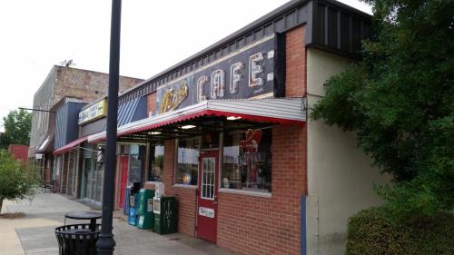 A brick cafe with a vintage sign, featuring outdoor seating and nearby storefronts in a small town setting.
