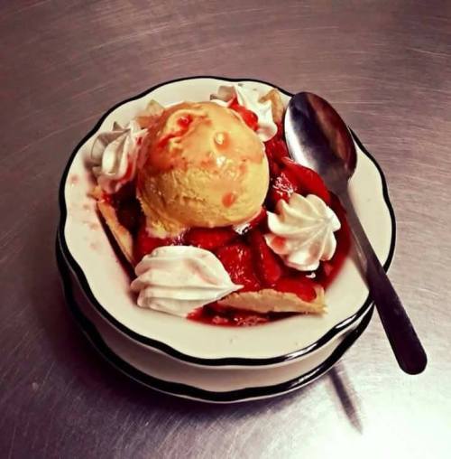 A dessert bowl with strawberries, vanilla ice cream, whipped cream, and a spoon on a metallic surface.