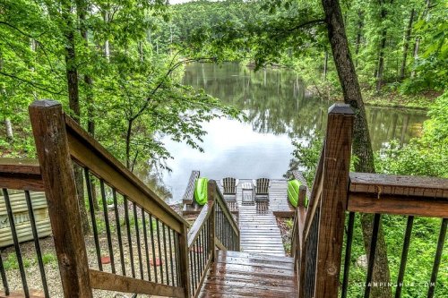 A wooden staircase leads down to a serene lake surrounded by lush greenery, with two chairs on the dock.