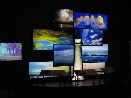 A display of various sky and ocean images surrounding a lighthouse model in a dark exhibition space.