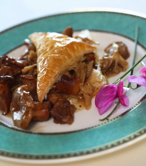 A plate featuring a golden pastry with mushrooms and a creamy sauce, garnished with purple orchids.