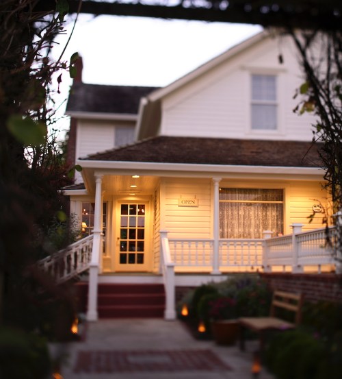 A charming white house with a welcoming porch, softly lit and marked "OPEN," surrounded by greenery and lanterns.