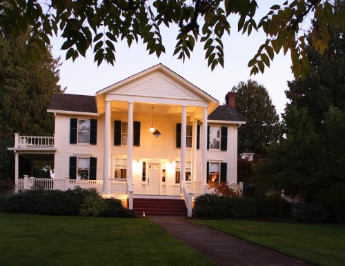 A charming white house with columns, surrounded by greenery, illuminated at dusk.