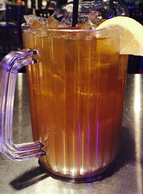 A clear pitcher filled with iced tea, ice cubes, and a lemon slice on the rim, sitting on a metallic surface.