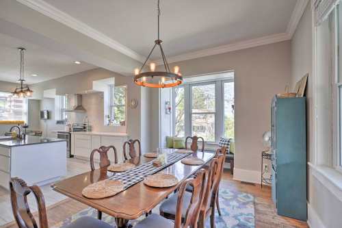 A bright dining area with a wooden table, chairs, and large windows overlooking greenery. Modern kitchen in the background.