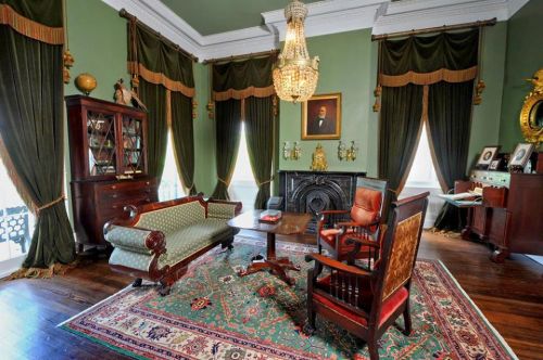 Elegant room with green walls, vintage furniture, a chandelier, and rich drapes, featuring a large area rug.