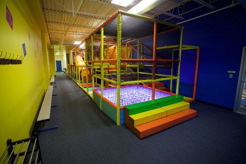 Colorful indoor play area with climbing structures, nets, and a soft landing zone, set against bright yellow and blue walls.