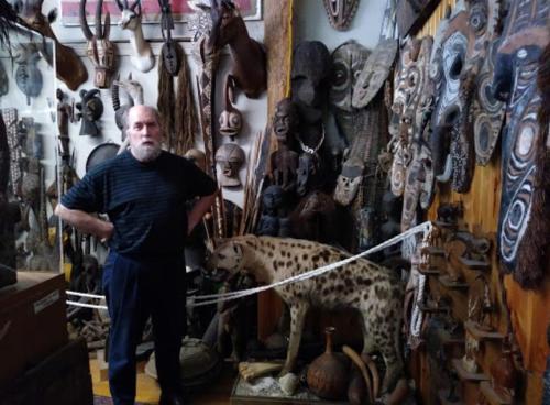 A man stands in a room filled with various tribal artifacts and masks, alongside a lifelike model of a hyena.