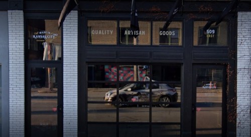Exterior view of a storefront with large windows displaying "Quality Artisan Goods" and a reflection of a car.
