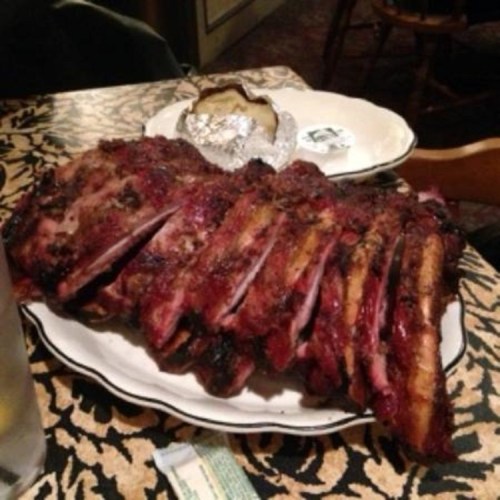A plate of grilled ribs served with a baked potato, set on a patterned tablecloth in a restaurant.