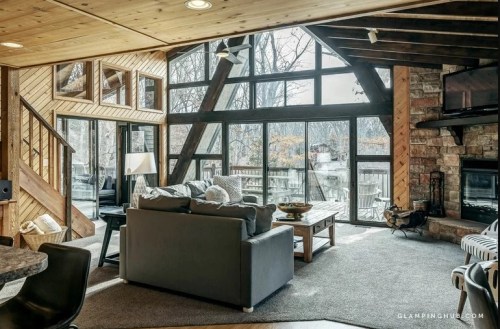 Cozy living room with large windows, wooden accents, and a stone fireplace, overlooking a serene outdoor view.