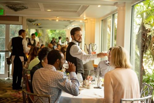 A waiter serves drinks to guests at a lively restaurant with large windows and tropical plants in the background.