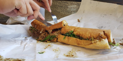 A person cuts into a large sandwich filled with meat and greens, resting on crumpled white paper.