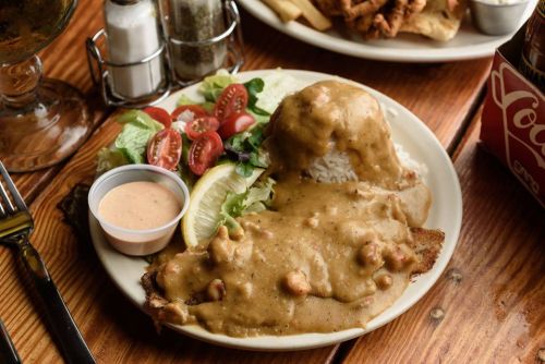 A plate of fried fish topped with creamy sauce, served with rice, salad, cherry tomatoes, and a lemon wedge.