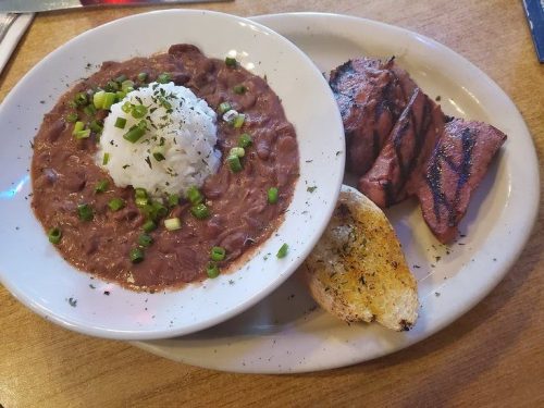 A plate featuring red beans and rice topped with green onions, grilled meat, and a slice of toasted garlic bread.