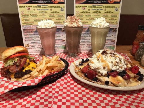A burger with bacon and cheese, fries, and a plate of waffles topped with fruit and whipped cream, alongside milkshakes.