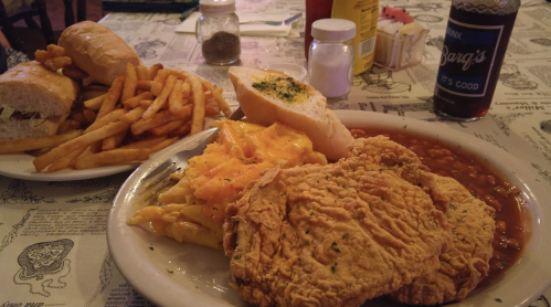 A plate of fried chicken, macaroni and cheese, baked beans, and garlic bread, with sandwiches and fries in the background.