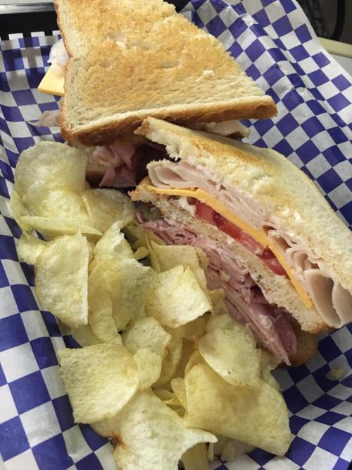 A sandwich with layers of deli meats, cheese, and tomato, served with a side of potato chips in a checkered basket.