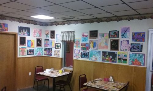 A cozy restaurant interior featuring a wall adorned with colorful children's artwork and simple wooden tables and chairs.