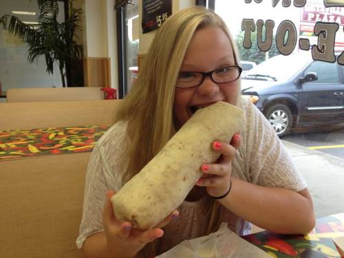 A smiling girl with long hair holds a large burrito in a casual dining setting.