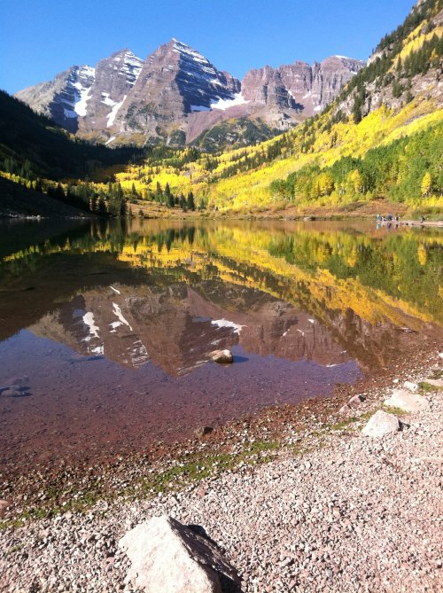A serene lake reflects colorful autumn trees and snow-capped mountains under a clear blue sky.