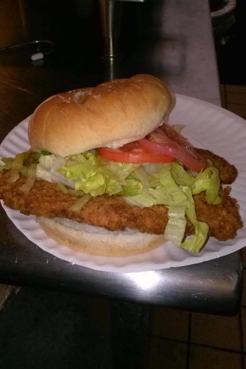 A crispy fried chicken sandwich with lettuce, tomato, and mayonnaise on a soft bun, served on a paper plate.