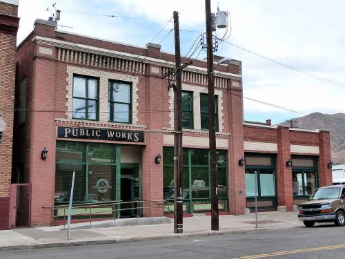 Historic brick building with large windows, featuring a "Public Works" sign, set on a street with utility poles.